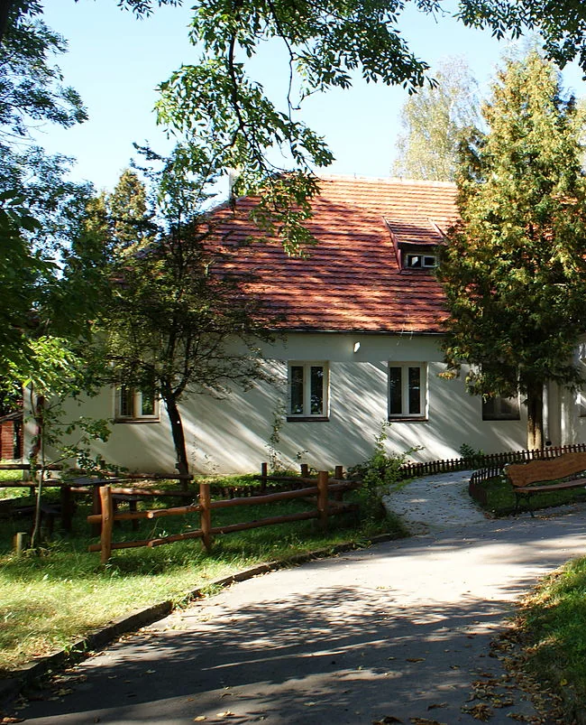 Fragment budynku ośrodka MONAR w Głoskowie z czerwonym dachem, w otoczeniu drzew.