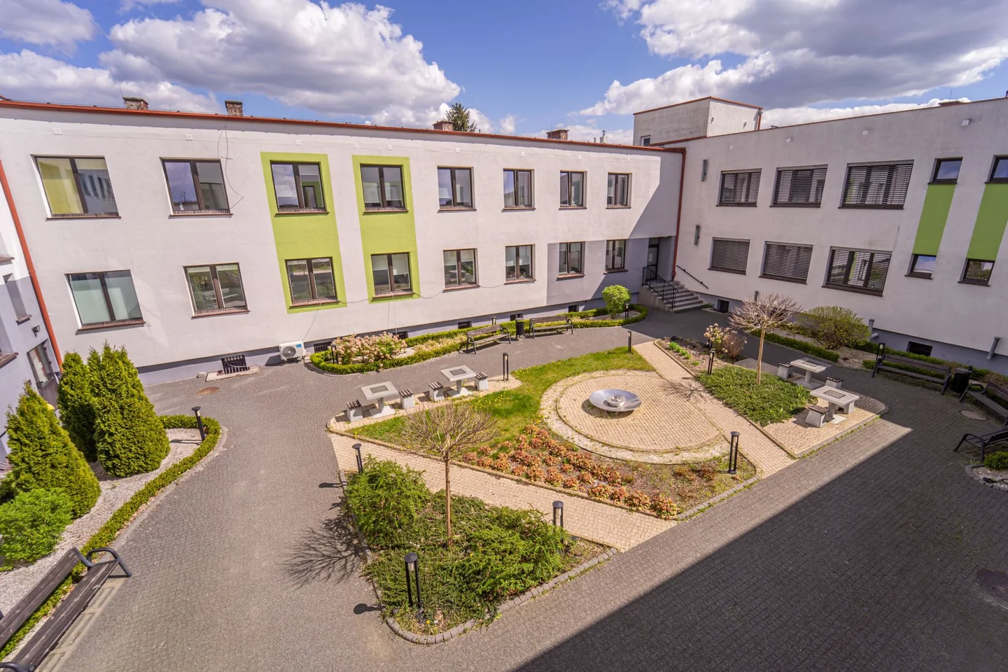 Dziedziniec pawilonu psychiatrycznego szpitala w Iławie, z ławkami i zielenią między skrzydłami budynku.
